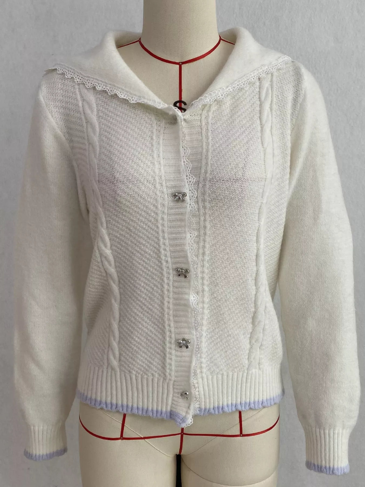 White cable-knit cardigan with lace trim, front on dress form.