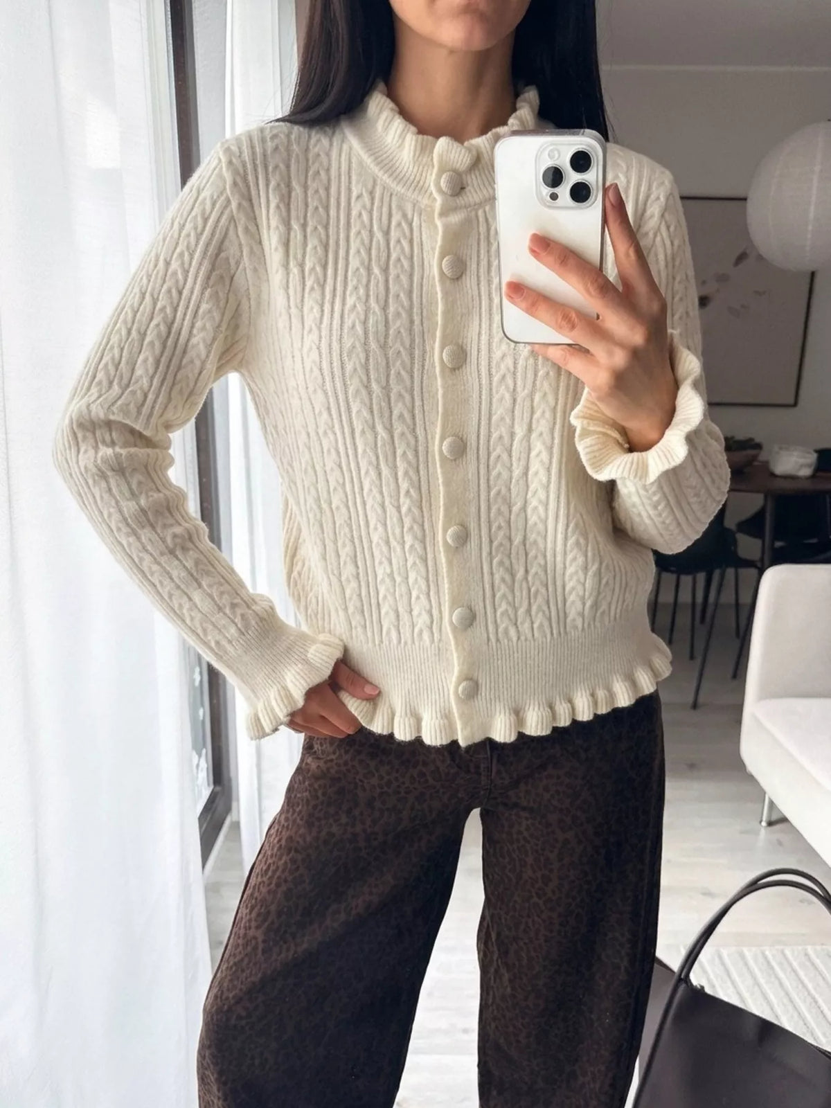 Ivory cable-knit ruffle-trim cardigan, front with hand at waist.